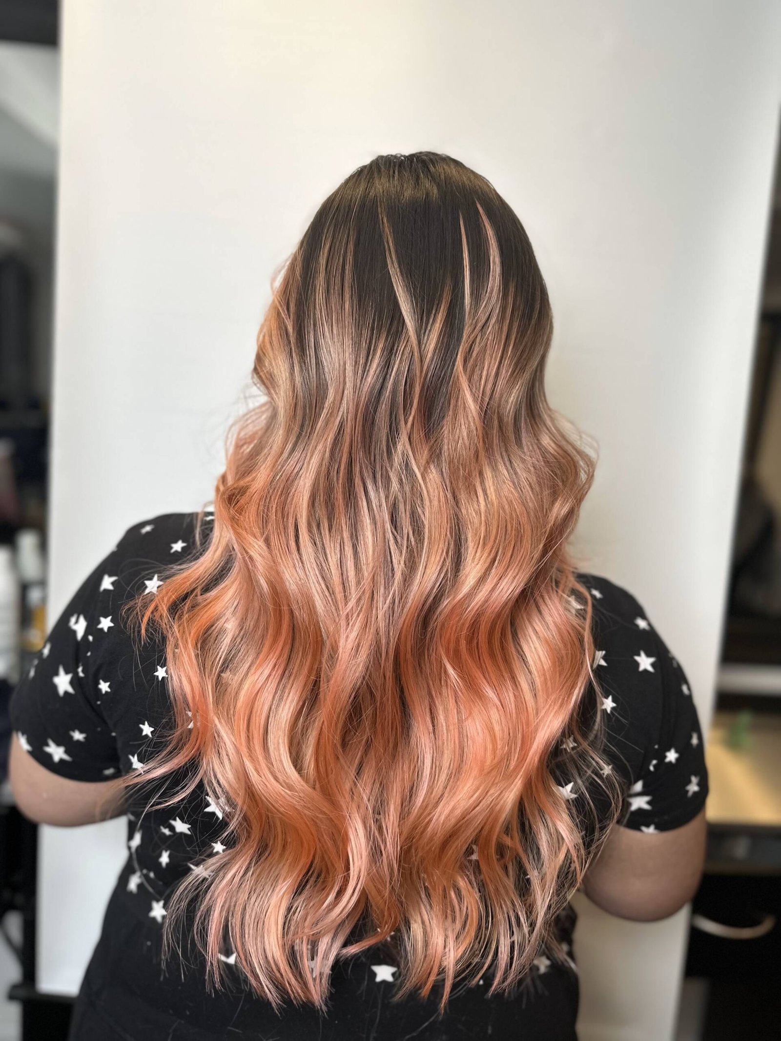A woman with long flowing hair in a rose gold ombre, transitioning from deep red at the roots to vibrant pink and finally into gold at the ends.